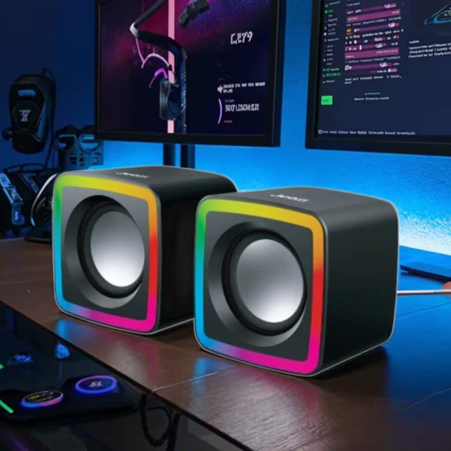 Pair of RGB speakers on a desk with computer monitors in the background