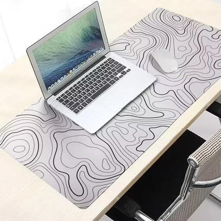 A large desk mat with a black topographic contour line pattern on a white background, styled on a light wood desk with a silver laptop and a white mouse.