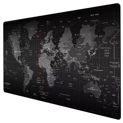 A large rectangular world map print or desk mat shown at an angle on a plain white background. The map is predominantly black and dark gray with lighter gray country outlines and text, plus a few small red line accents.